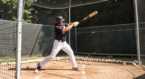 An adult player taking batting practice in a cage, mid-swing, with softballs scattered in the netting behind.