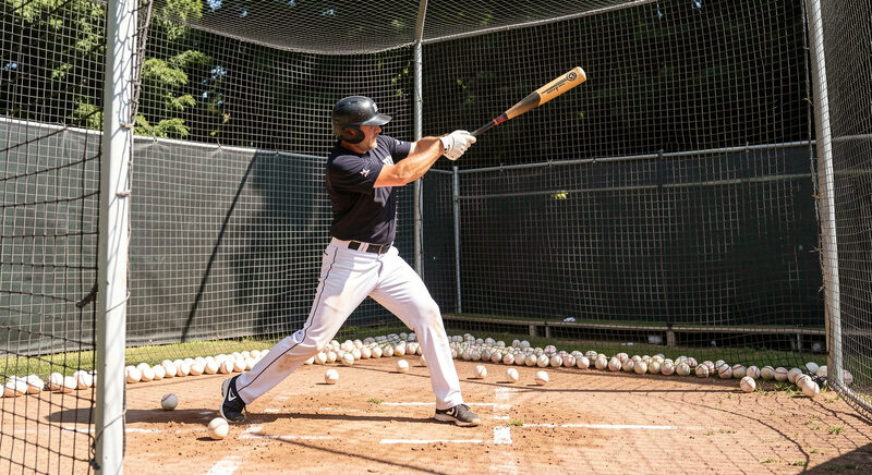 An adult player taking batting practice in a cage, mid-swing, with softballs scattered in the netting behind.
